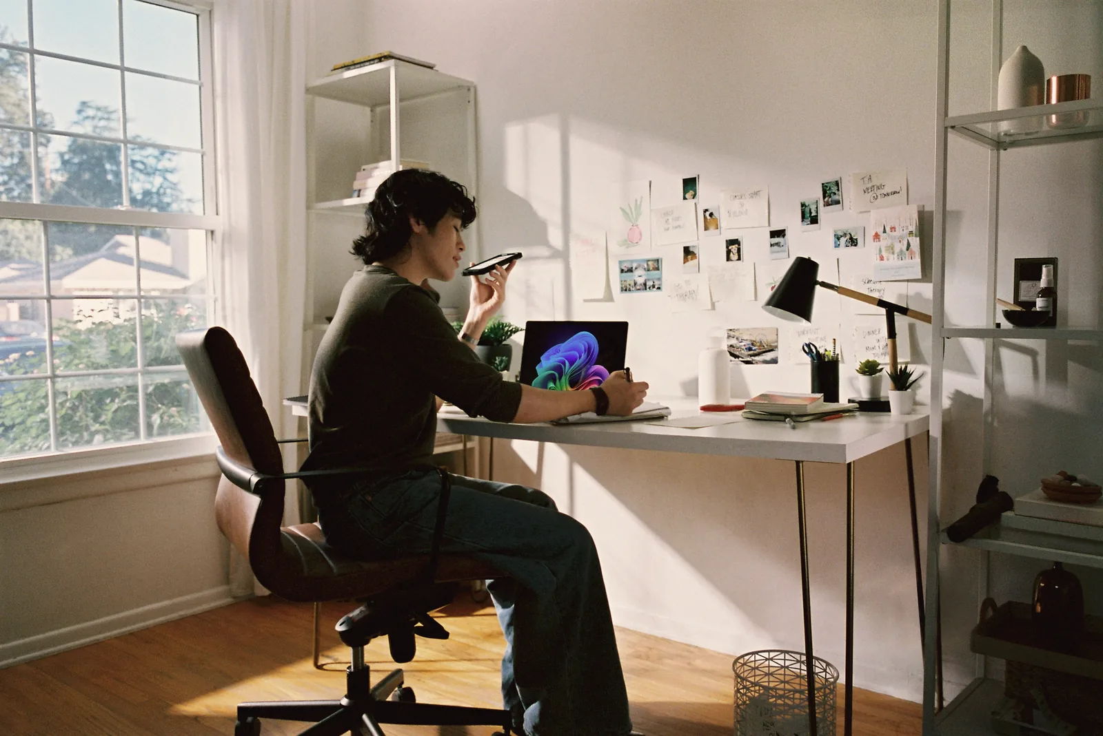 A woman on a phone call at her desk by the window, a wall of pinned photos behind her
